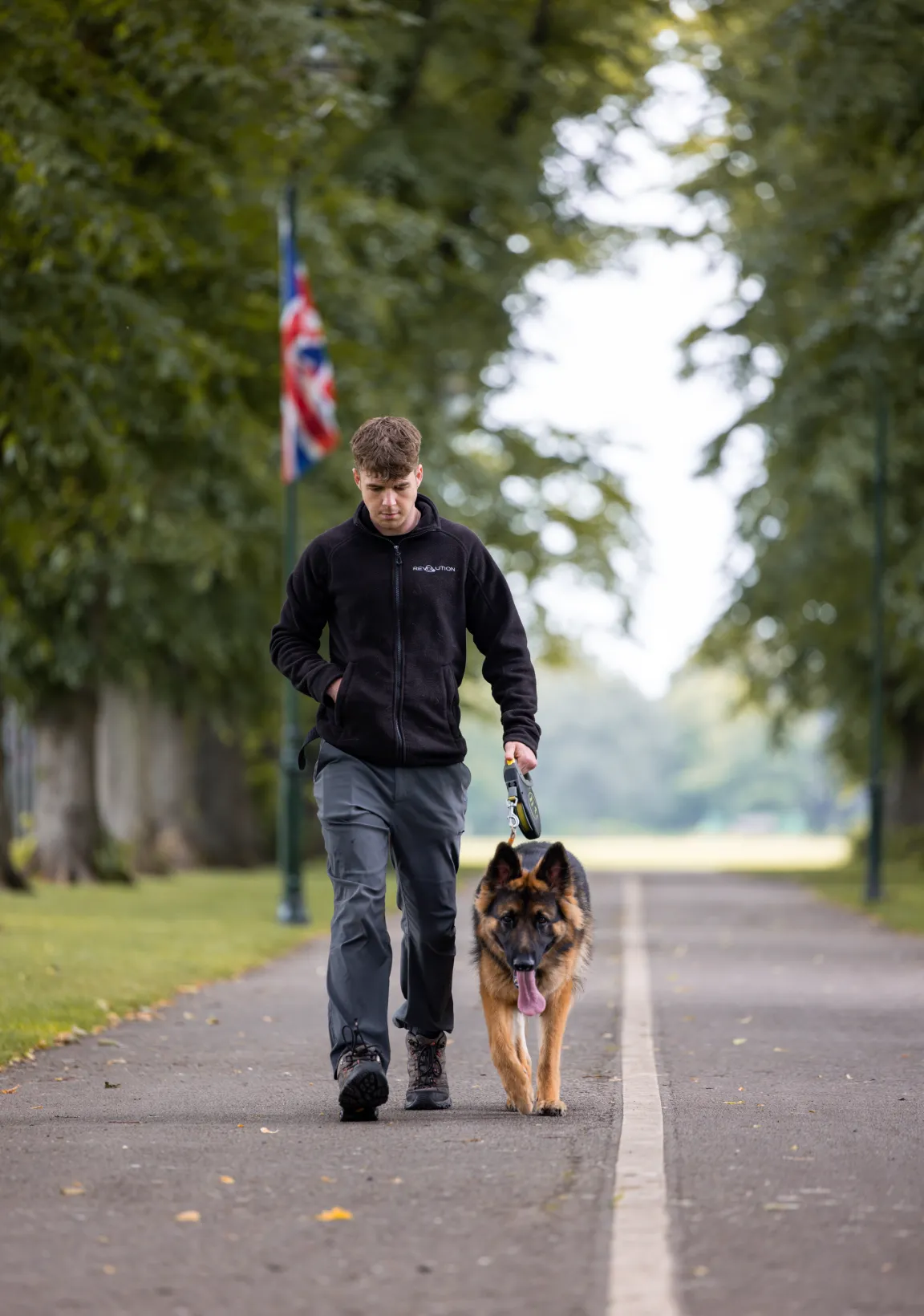 A trainer walking a German Shepherd to heel down a tree-lined path