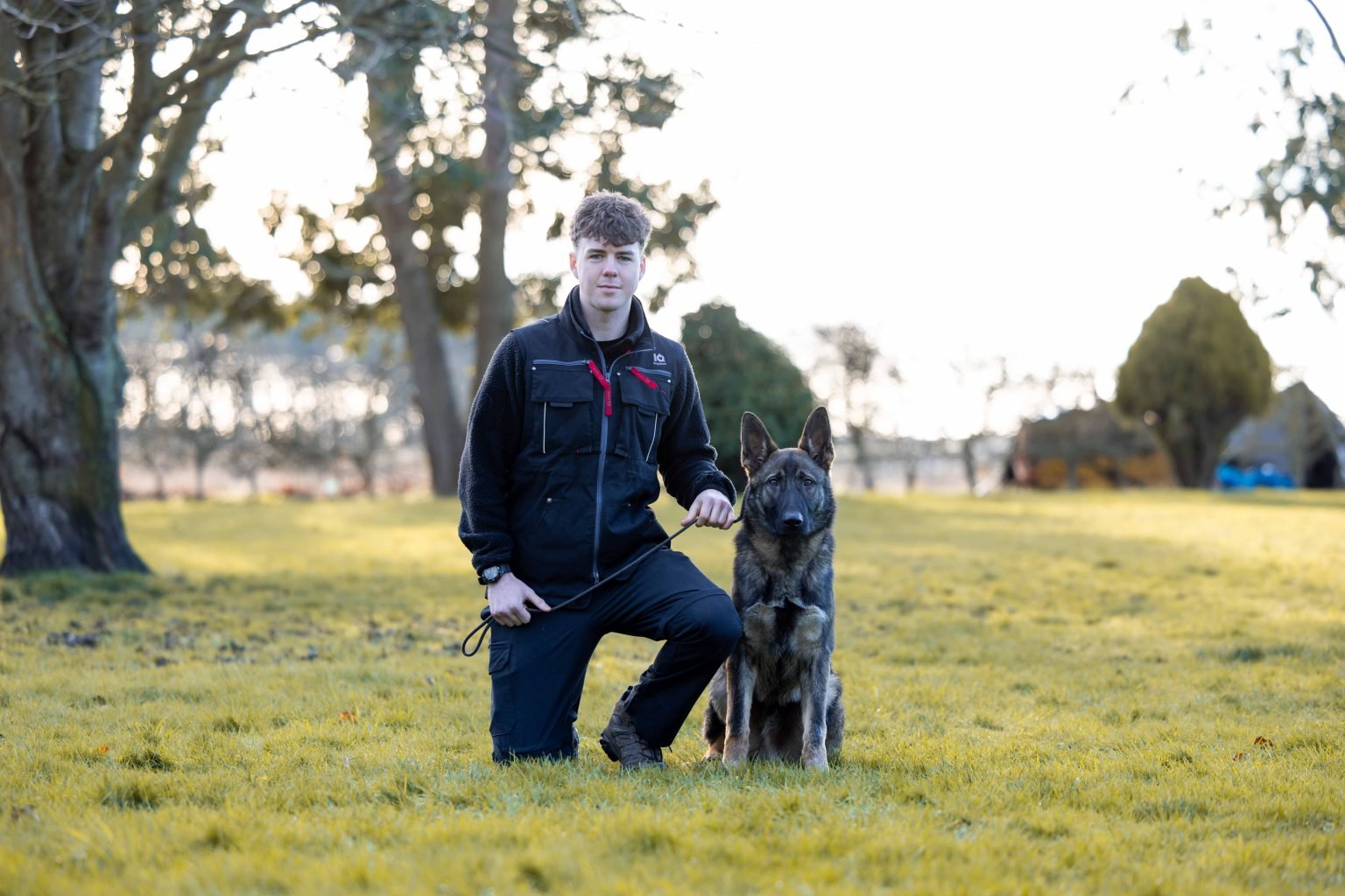Jhai Herrgesell kneeling beside a German Shepherd in a field
