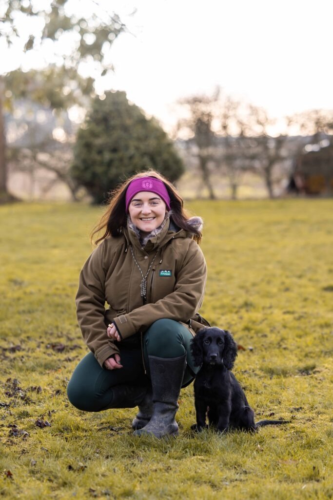 Nel Forth crouching in a field beside her black spaniel, Pepper